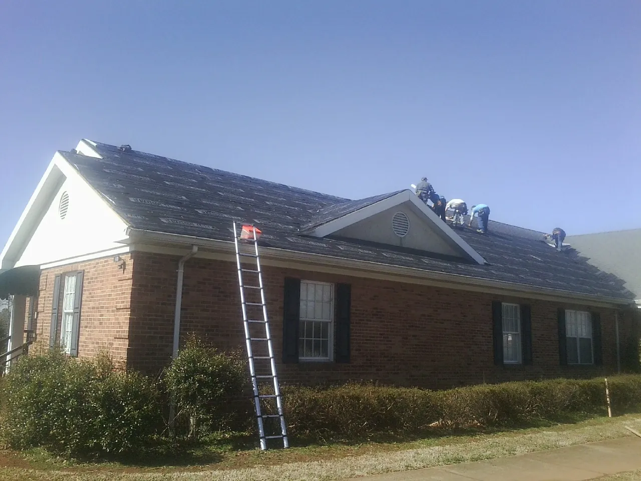 Crew tearing shingles off a brick ranch roof with a ladder set — Emergency Roof Repair work by our crew in Longford