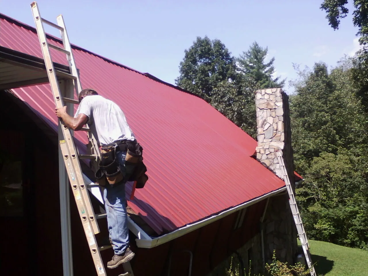 Roofer on a ladder beside a red metal roof and stone chimney — Soffit & Fascia Repair by Roofing Brothers in Longford